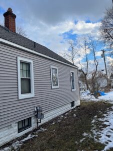 side view of a gray house with chimney and window, snow on ground and bare trees in background.