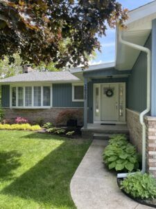 Front entrance of a greenish-gray house with wreath on door, walkway and landscaped plants
